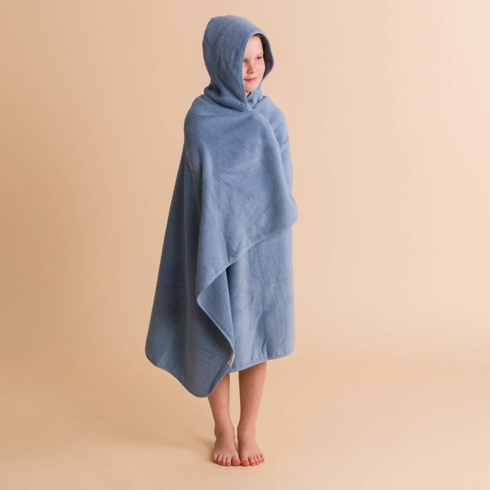 A child standing barefoot and wrapped in a dusty blue cotton terry hooded towel against a solid beige studio background.