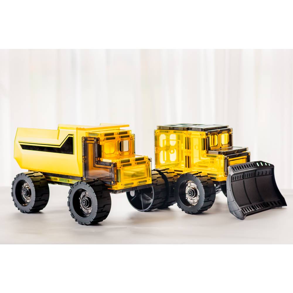 Two yellow magnetic construction vehicles, a dump truck and a bulldozer, displayed side by side on a light gray surface with a soft white curtain background.