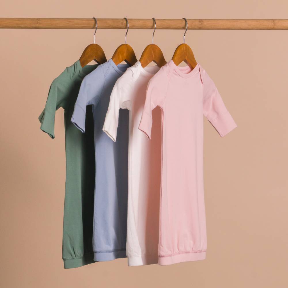 Four children's cotton night gowns in green, blue, white, and pink hanging on a rack against a beige background.