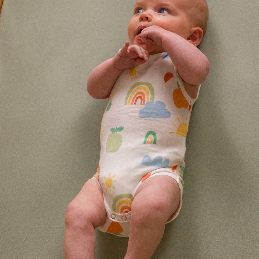 Baby wearing a colorful bodysuit with rainbow and cloud patterns on a plain background