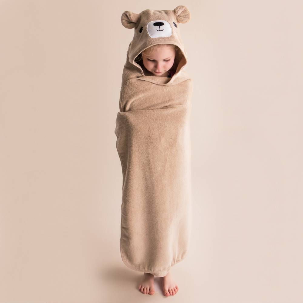 A baby wrapped in a plush terry cotton tan hooded towel with bear ears and a bear face on the hood, standing barefoot against a solid beige background.