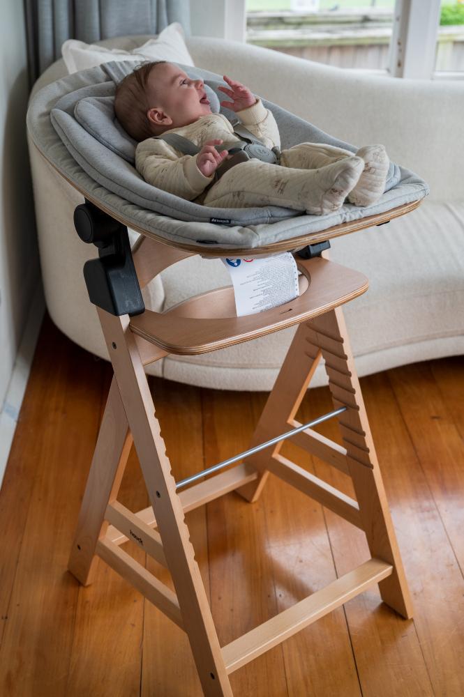 Baby in a Hauck high chair with Bouncer attached with a wooden frame on a wooden floor.