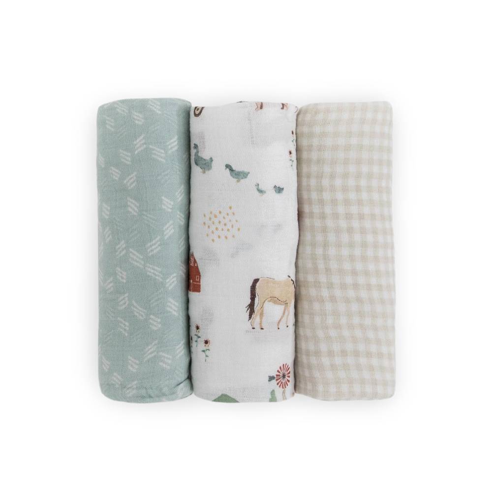 Three rolled muslin swaddle blankets standing upright on a white background. The left blanket is sage green with white leaf patterns.
