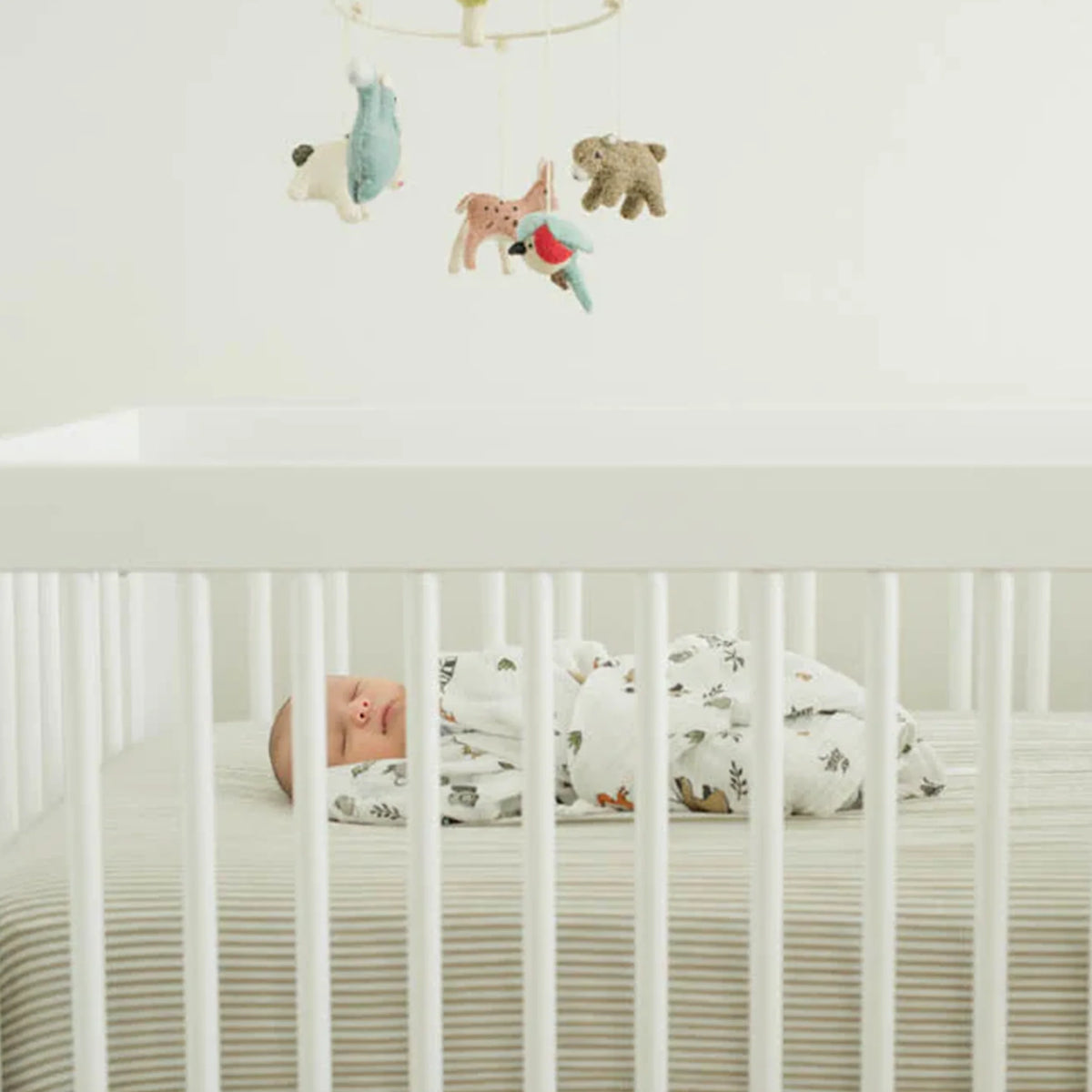 Baby sleeping in a white cot on a grey striped blanket with colorful animal mobile above