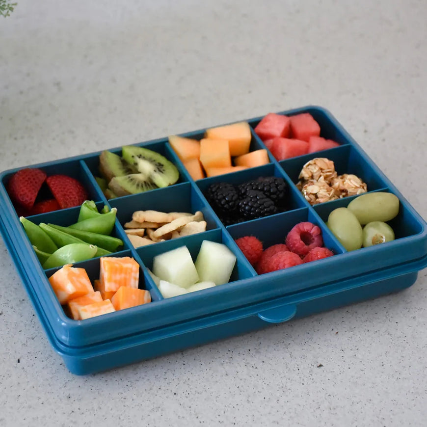 A blue compartment lunch box filled with fruit, vegetables, crackers, and granola, photographed on a light speckled countertop.
