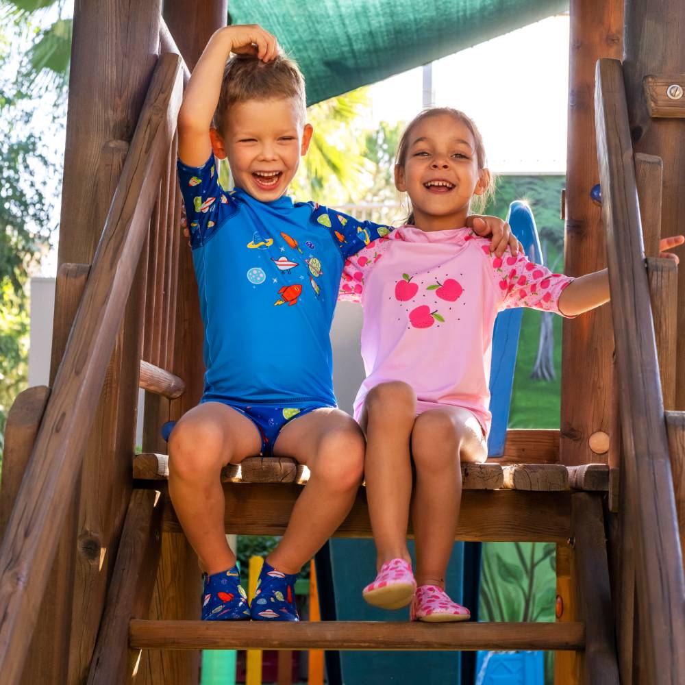 Two children sitting on wooden playground stairs wearing matching swim shirts and shorts sets. The boy wears a blue space-themed swim shirt with yellow, red, and blue graphics, and the girl wears a pink swim shirt with red apple graphics. They are wearing matching slipfree water shoes 