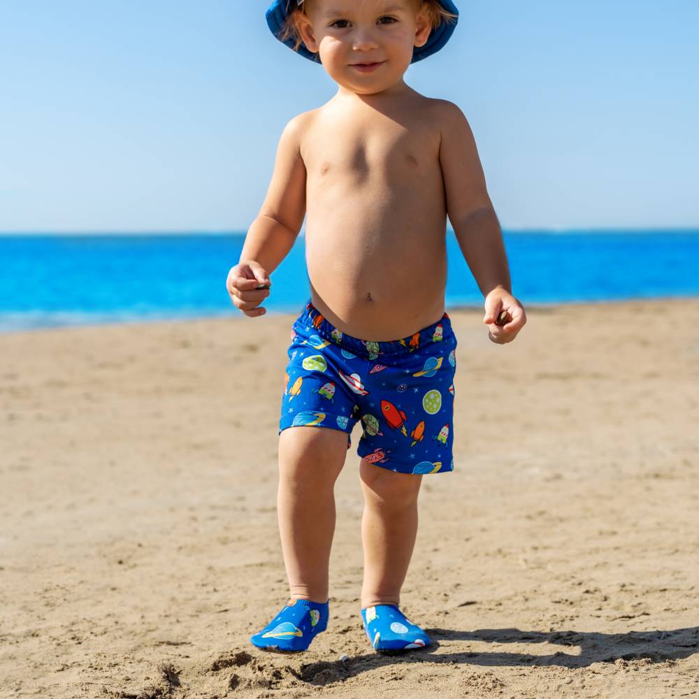 A toddler standing on a sandy beach wearing blue swim trunks with a colorful fish pattern, a blue sun hat, and blue slipfree water shoes, with the ocean and blue sky in the background.