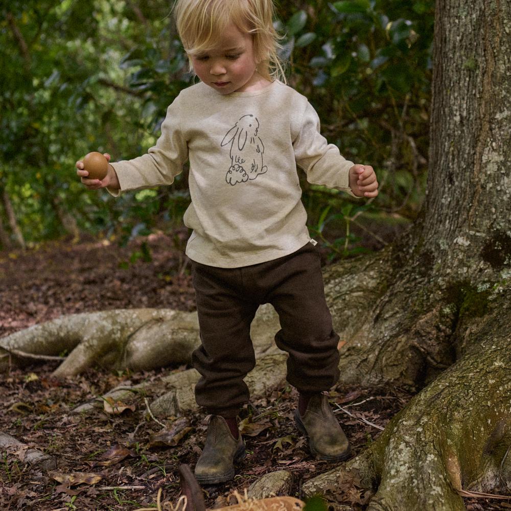 Child in a forest setting wearing a beige long-sleeve shirt with a bunny design and brown pants.
