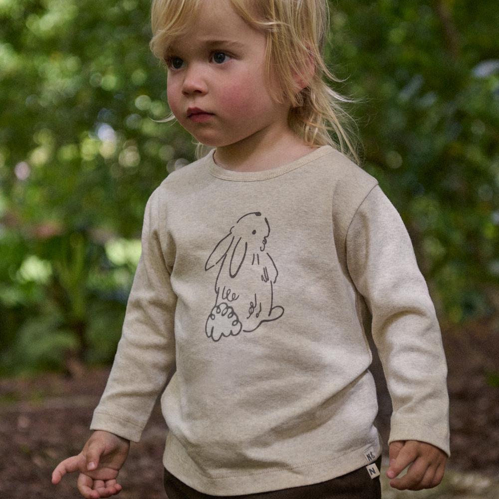 Child wearing a beige long sleeve shirt with a brown bunny design in an outdoor setting 