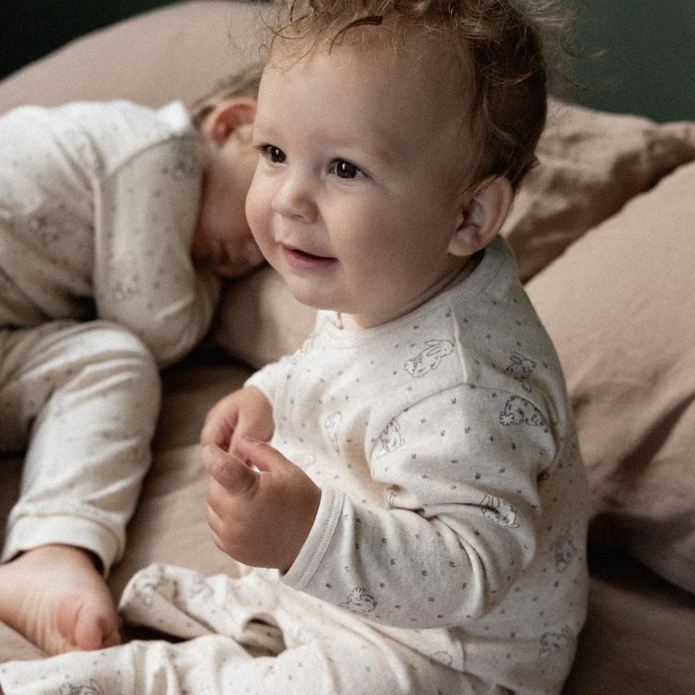 Two babies in matching beige and brown pyjamas with bunny pattern sitting on a bed with brown linen sheets