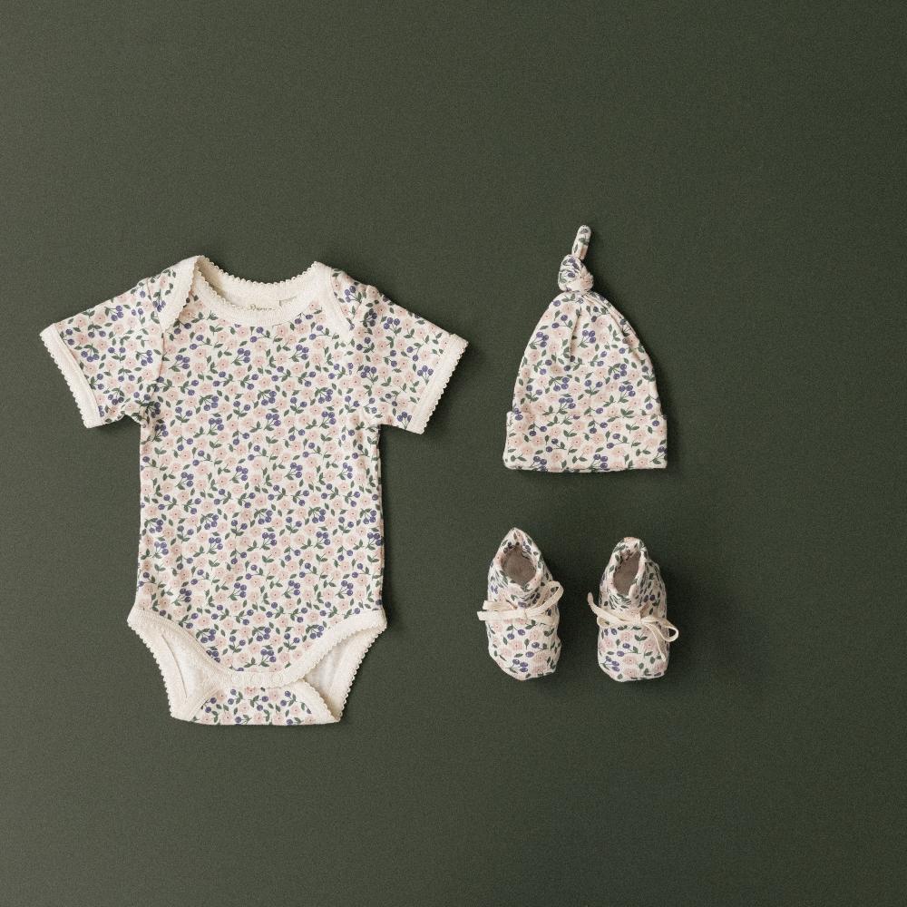 Cream and feminine floral Baby clothing set with floral-patterned bodysuit, hat, and booties shown on a dark green background