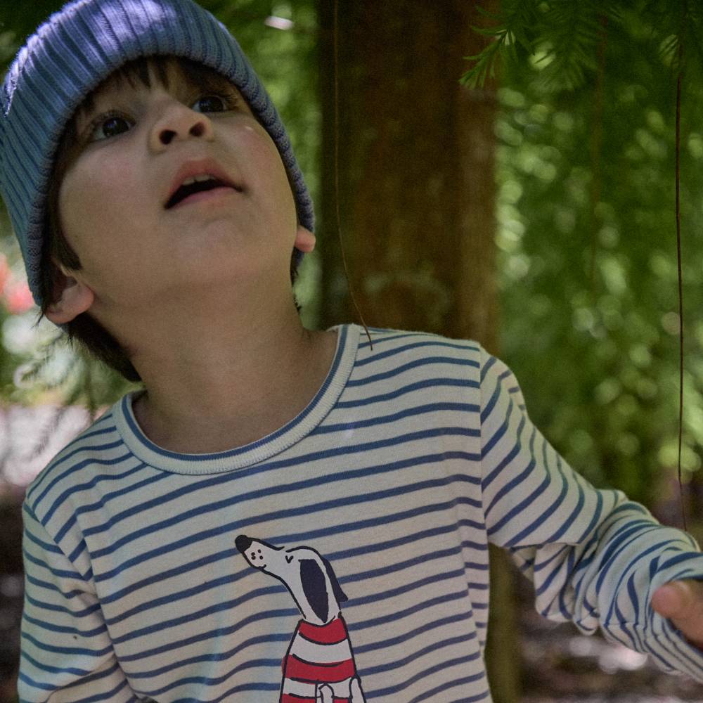 Child wearing a striped shirt with a dog design an an outdoor setting with a blue chunky knit beanie
