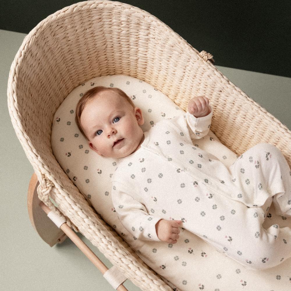 Baby wearing a cream baby footed onesie with a clover and pink pattern lying in a wicker Moses basket.