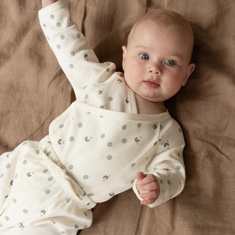 Baby wearing a white outfit including kimono bodysuit and footed leggings with green clover patterns lying on a brown surface