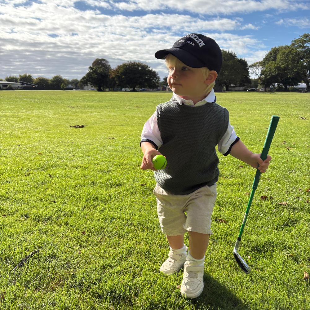 Young child in golf attire including a forest green woolen vest holding a golf club and ball on a grassy field.