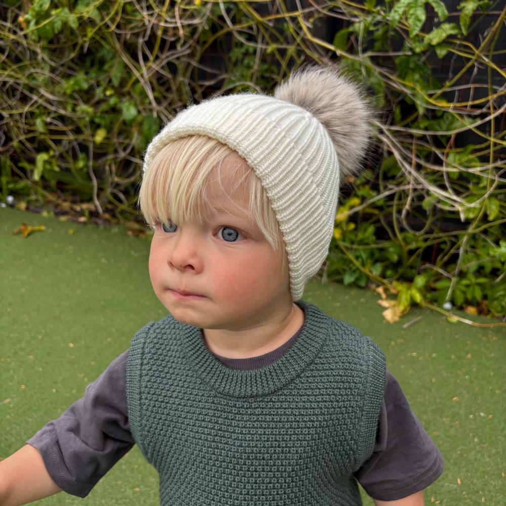 Child wearing a merino wool knit hat with a pom-pom outdoors on a green surface.