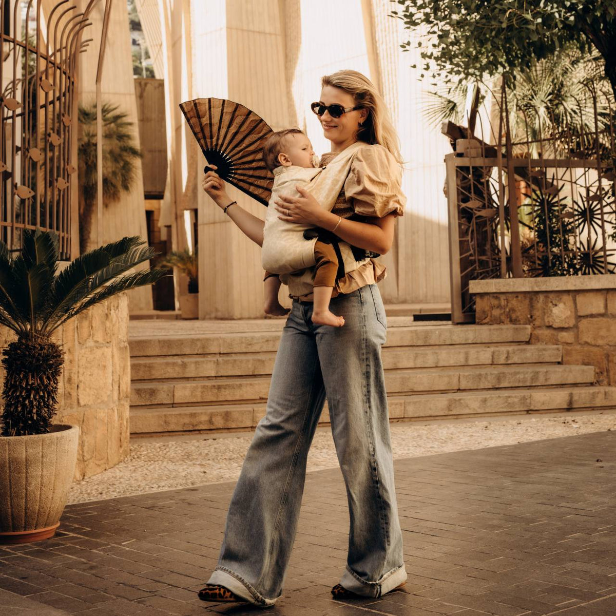 A woman walking down the street wearing her baby in a beige baby carrier on the front facing in. 