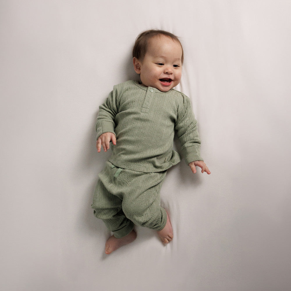 Baby lying on a mattress wearing a Flax Marl Woolbabe Merino Cotton Rib Henley Top and Pants. 
