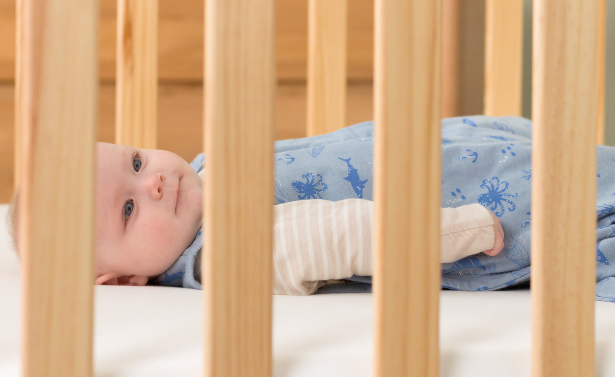 Baby lying in a woolbabe blue sleep sack in a crib with wooden bars