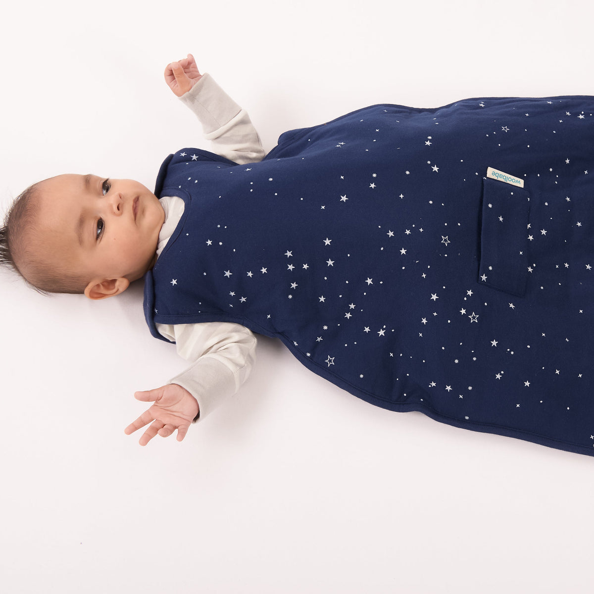 A baby lying on their back wearing a navy blue sleep bag with a white star pattern, photographed on a clean white background.