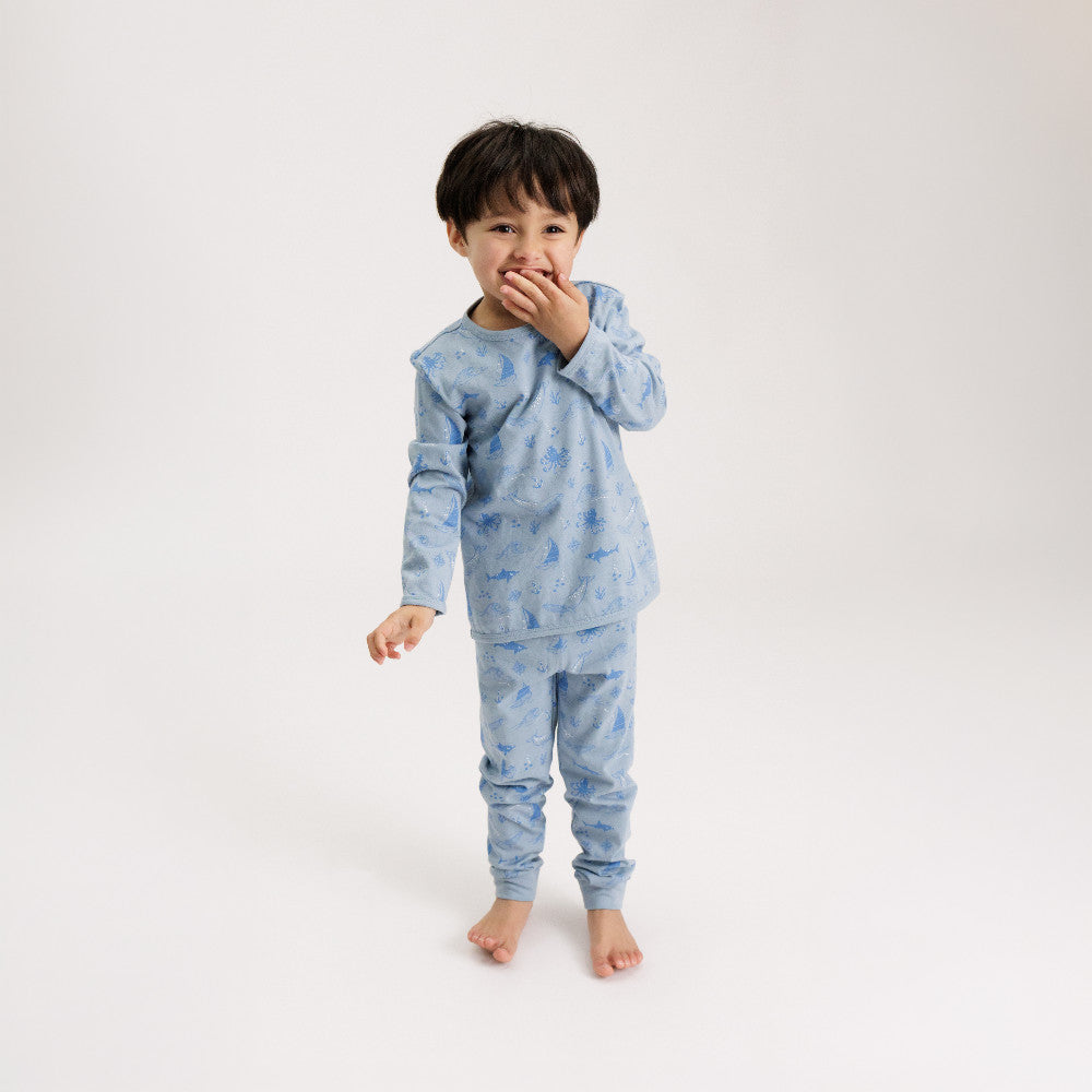 A young boy wearing a light blue long-sleeve pajama set with a darker blue pattern, standing against a plain white studio background.