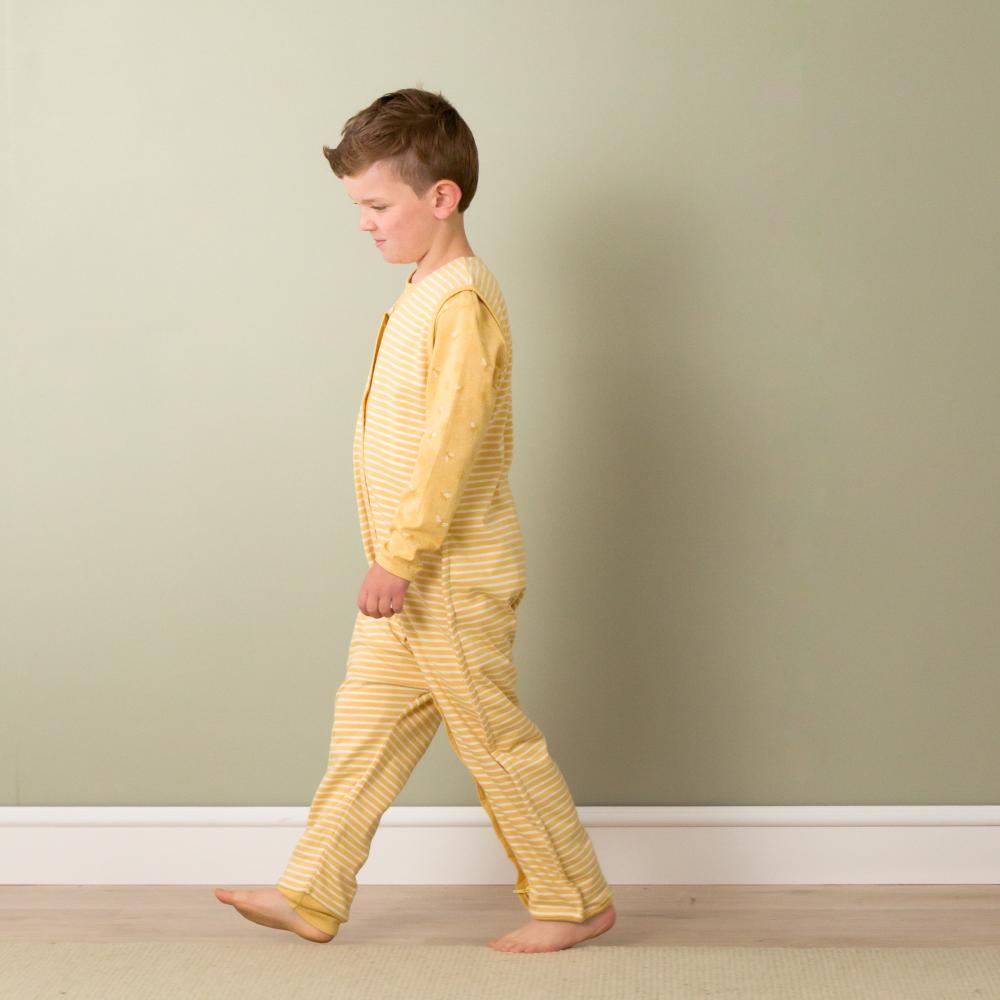 Child wearing yellow sleepsuit walking against a plain wall.