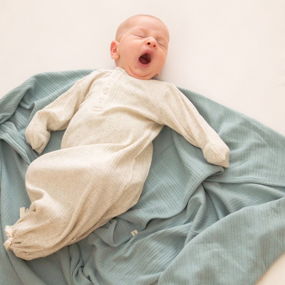 Newborn baby yawning dressed in a cream baby nightgown lying on a light blue woolbabe swaddle 