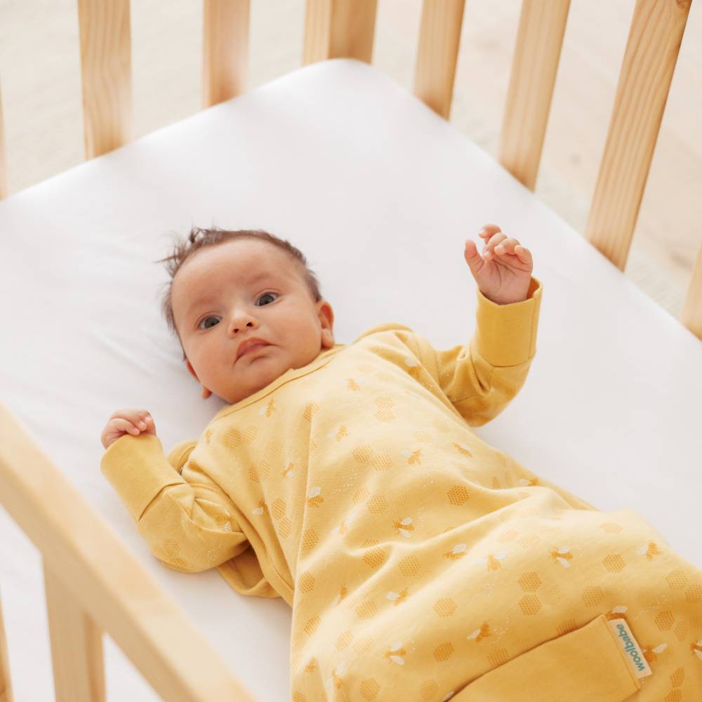Baby in a yellow onesie lying in a small size wood crib with white bedding