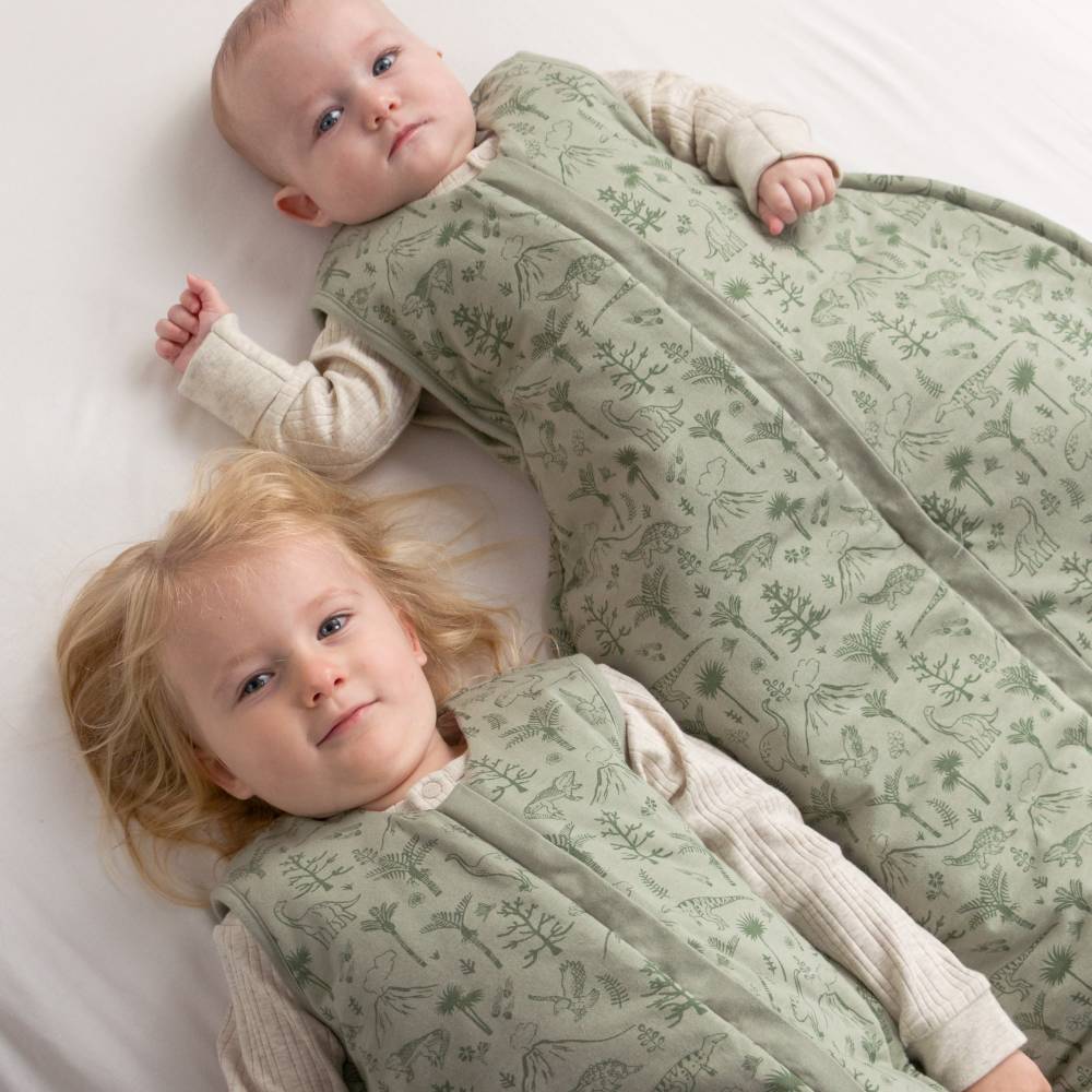 Two children wearing green dinosaur patterned sleep sacks lying on a white surface.