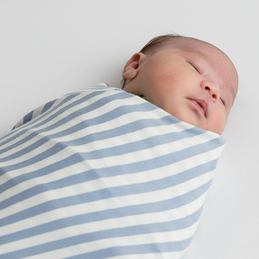 A sleeping newborn baby wrapped in a blue and white striped  woolbabe swaddle blanket, lying on a plain light gray background.