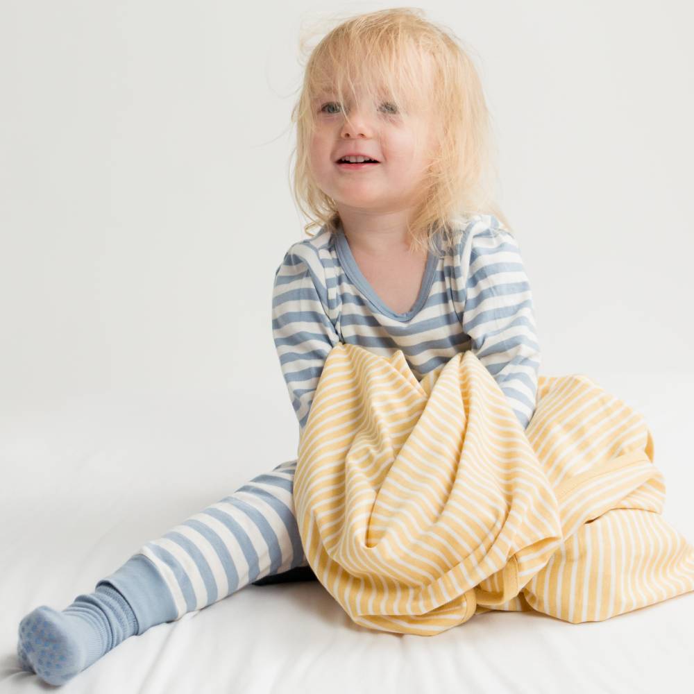 Child wearing blue striped woolbabe pajamas with a yellow blanket on a white background