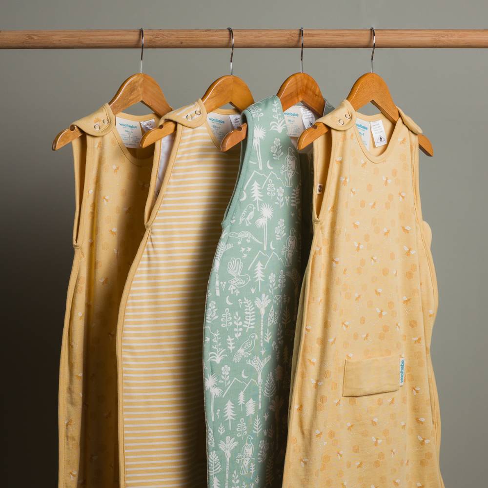 Four woolbabe baby wearable blanket sleeping bags hanging on a rack against a gray background