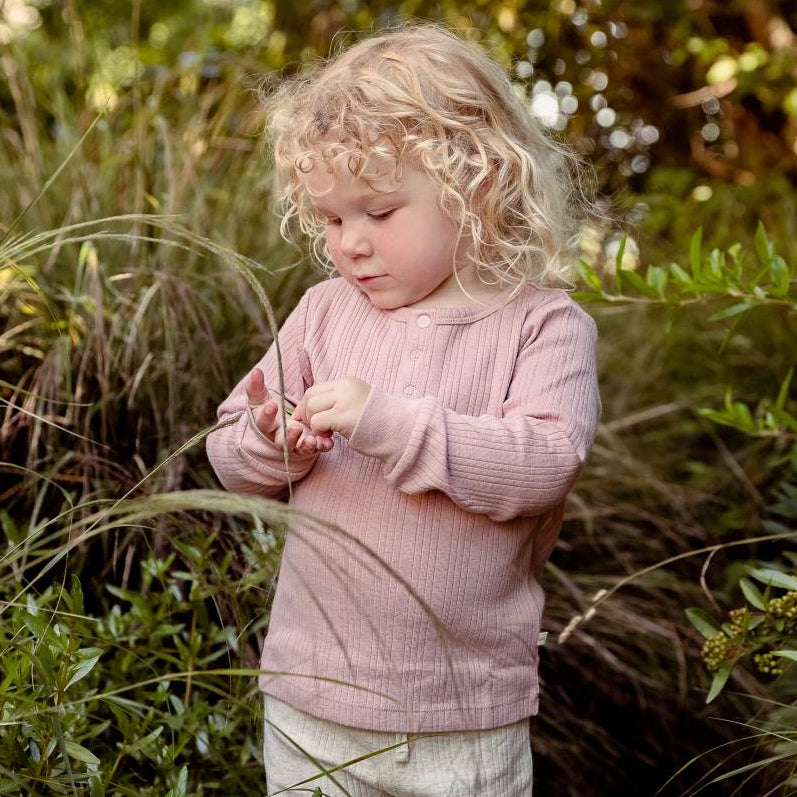 Child in a pink woolbabe top standing in a natural setting with greenery.