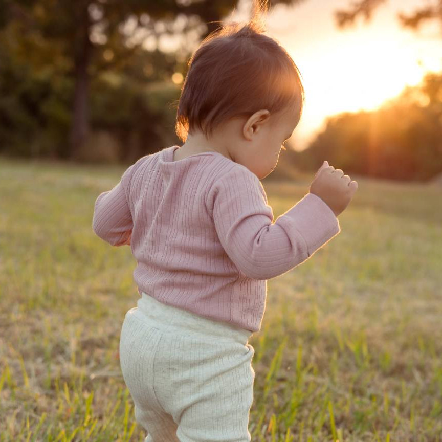 Child in a pink bodysuit and light pants standing in a grassy field with a sunset in the background