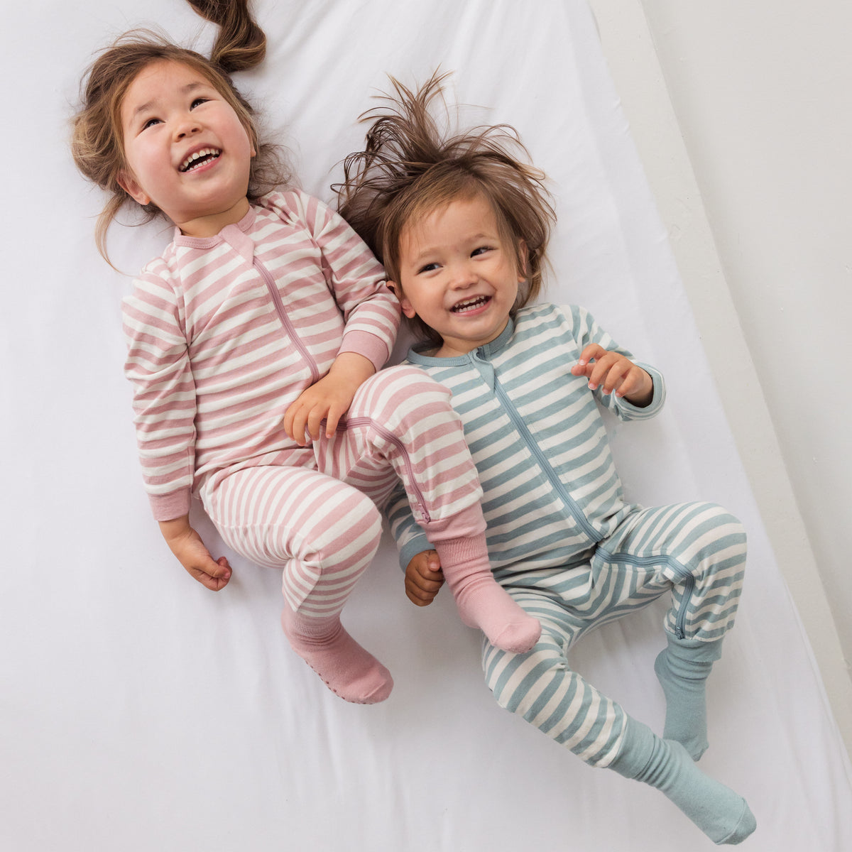 Two children wearing matching striped pajamas and woolbabe socks  lying on a white surface.