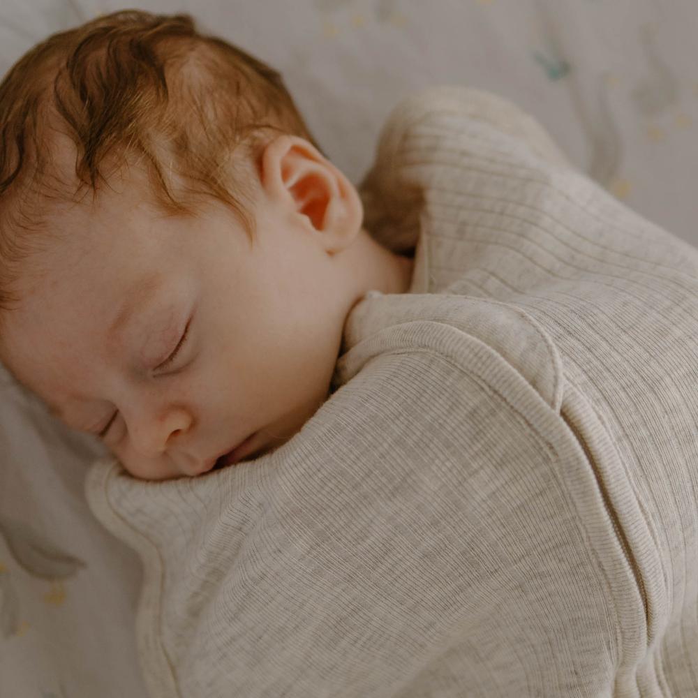 Baby sleeping peacefully wrapped in a beige woolbabe merino zip swaddle with a soft focus background