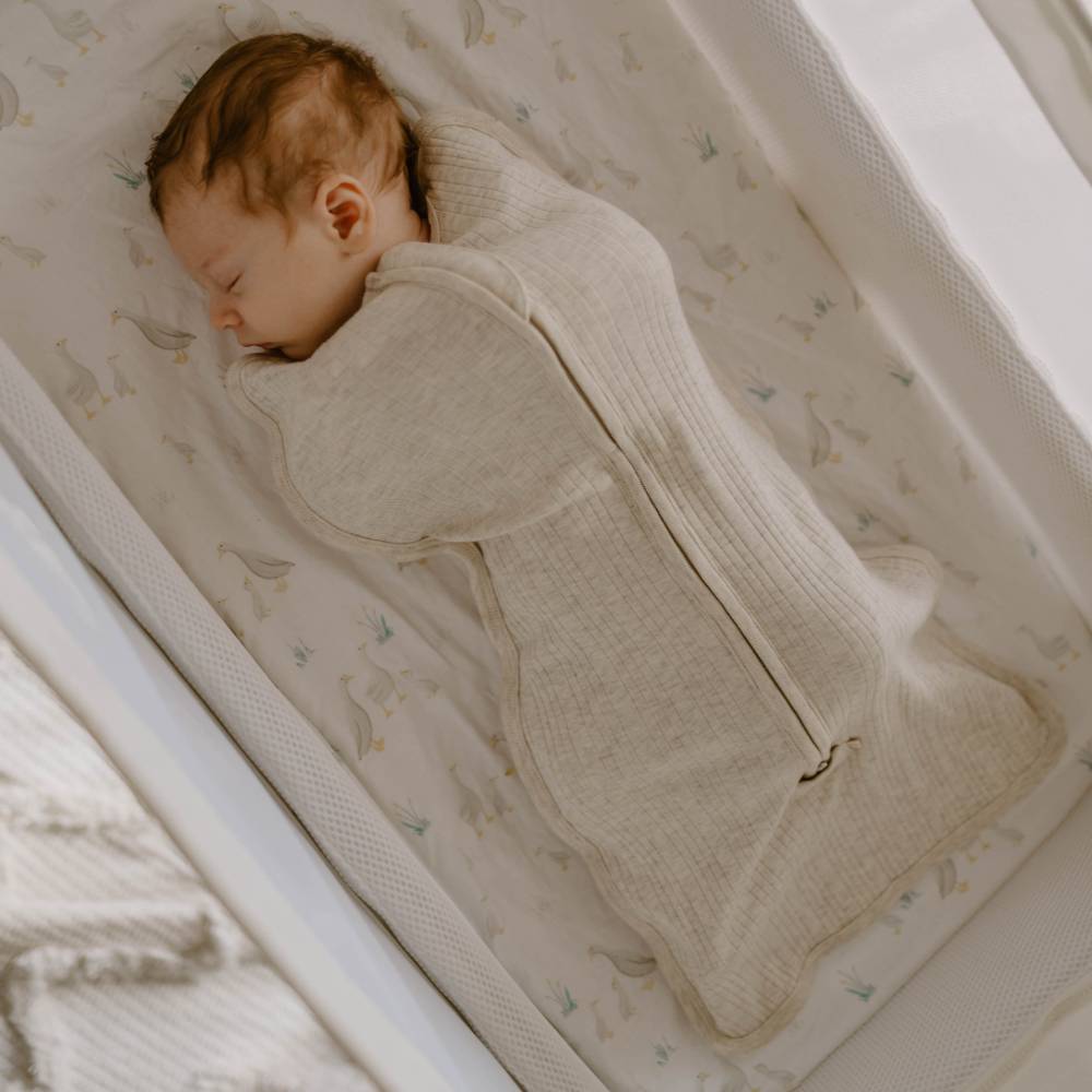 Newborn baby sleeping in a crib wearing a woolbabe zip up swaddle with their arms on chest