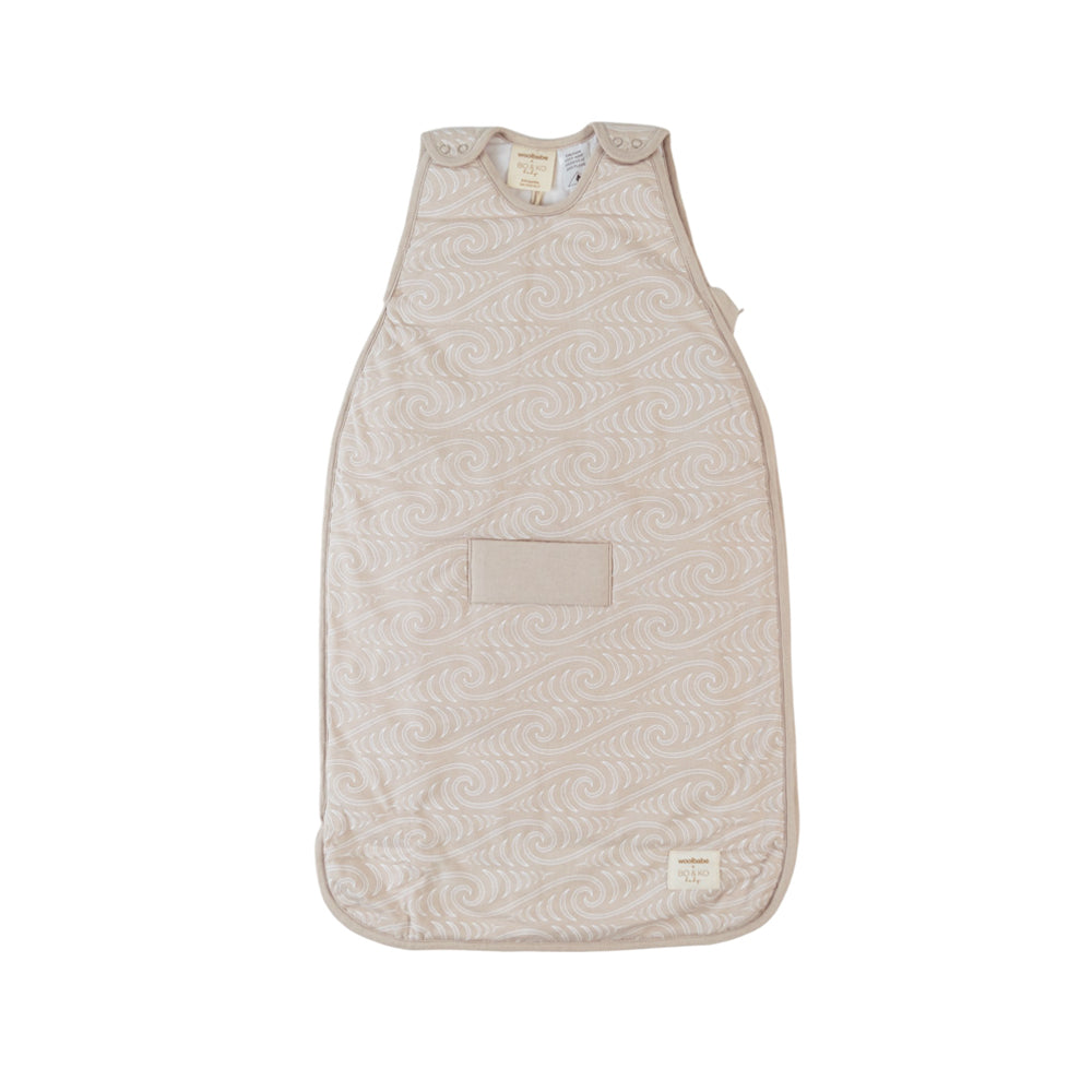 A baby sleep sack in a light beige color with a subtle Maori designed wavy pattern, featuring a front patch pocket and a small brand label near the bottom right corner.