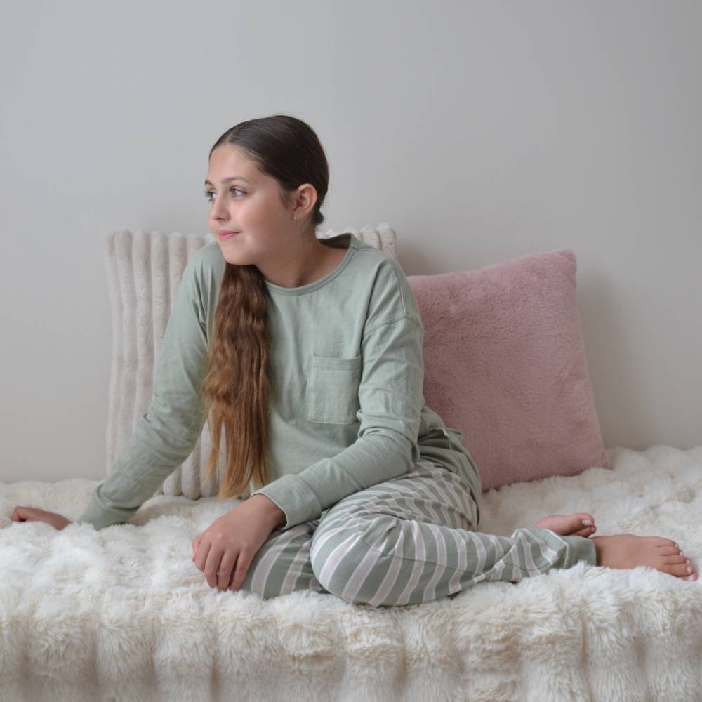 Teenage girl  wearing a light green long-sleeve top and striped pants sitting on a bed with pink pillows.
