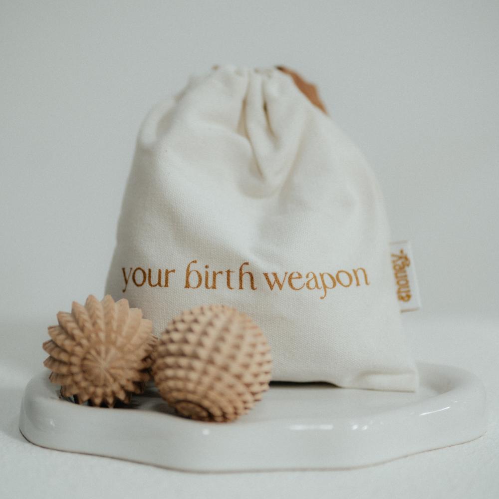 An off-white fabric drawstring pouch with the words 'your birth weapon' printed in brown, displayed on a white tray with two textured, spiky massage balls. 