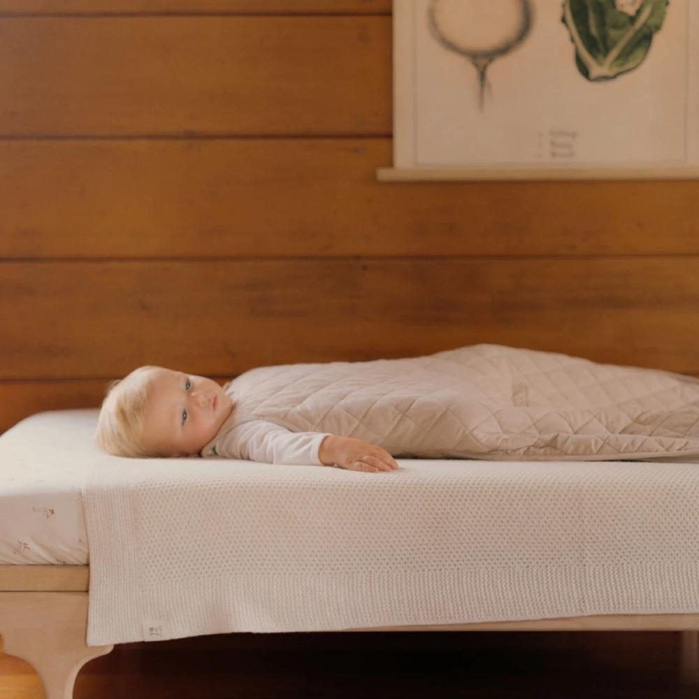 Baby lying on a bed with a wooden headboard and framed picture wearing a beige quilted sleeping bag.