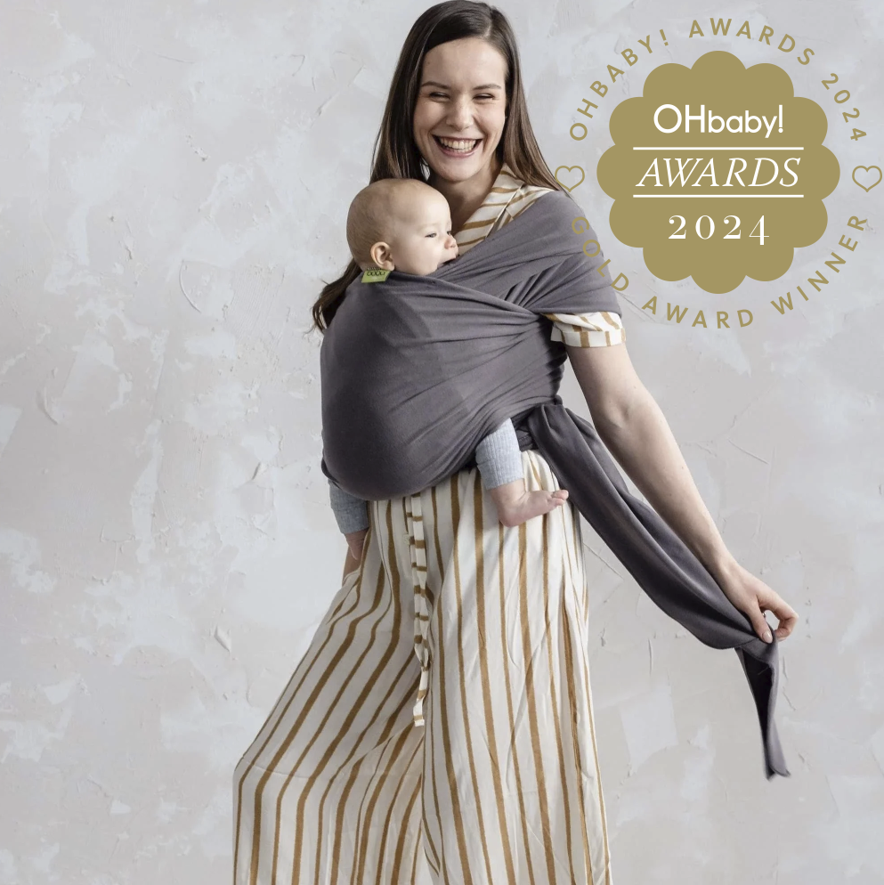 A woman wearing a cream and tan vertically striped dress carries a baby in a dark gray wrap-style baby carrier. The photo is set against a textured light gray wall, with an 'Ohbaby! Awards 2024 Award Winner' badge in the top right corner.