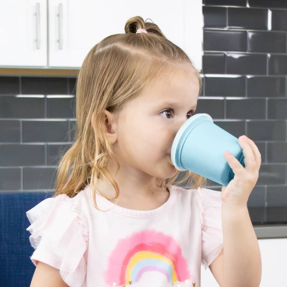A young girl with blonde hair drinks from a light blue plastic cup, wearing a pink t-shirt with a rainbow graphic, set against a kitchen background with dark gray subway tiles.