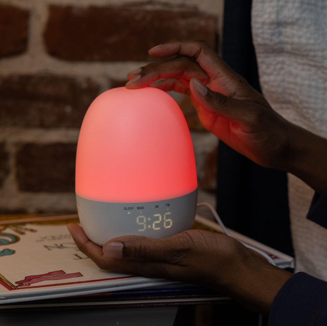 Person holding a glowing sleep trainer clock with digital display against a brick wall.