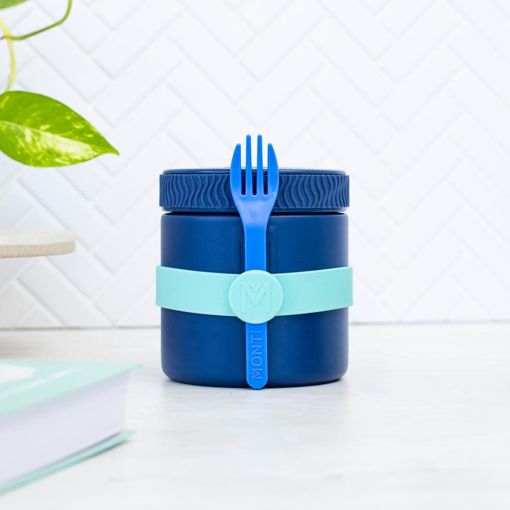A dark blue Montii food storage container with a matching blue lid and a light blue silicone band, shown on a white surface with a tiled background and a plant leaf in the corner.