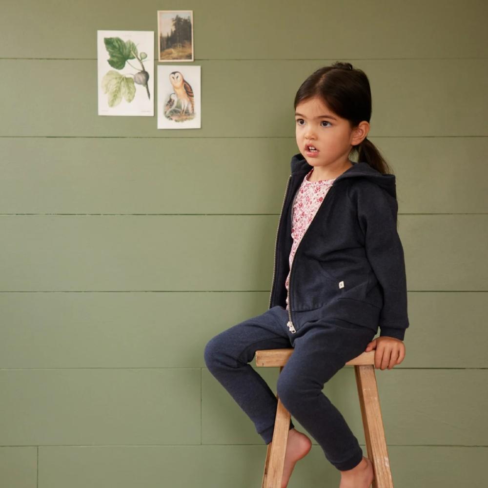 Young girl sitting on a stool against a green wall with nature-themed posters wearing blue trackpants and navy zip up sweater 