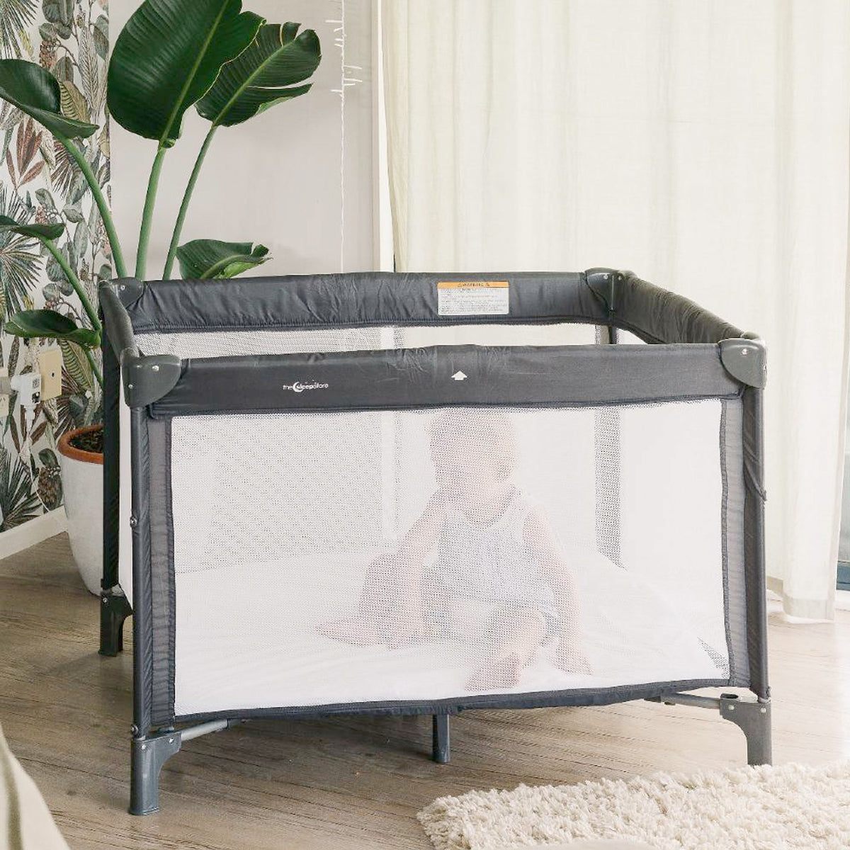 Child in a gray portable travel cot with mesh side in a room with plants and curtains
