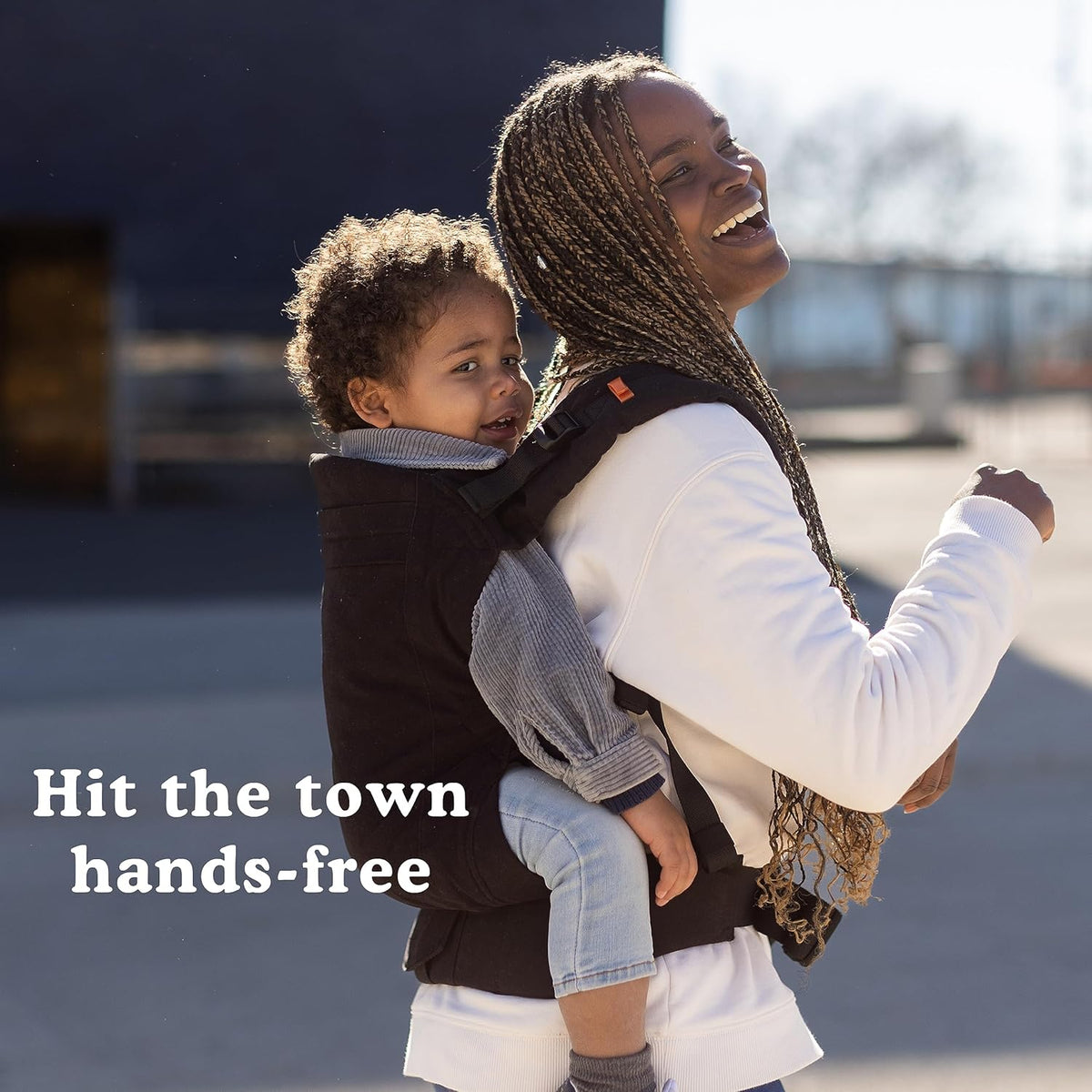 Woman carrying a child in a Beco Toddler carrier with text 'Hit the town hands-free' on a blurred background